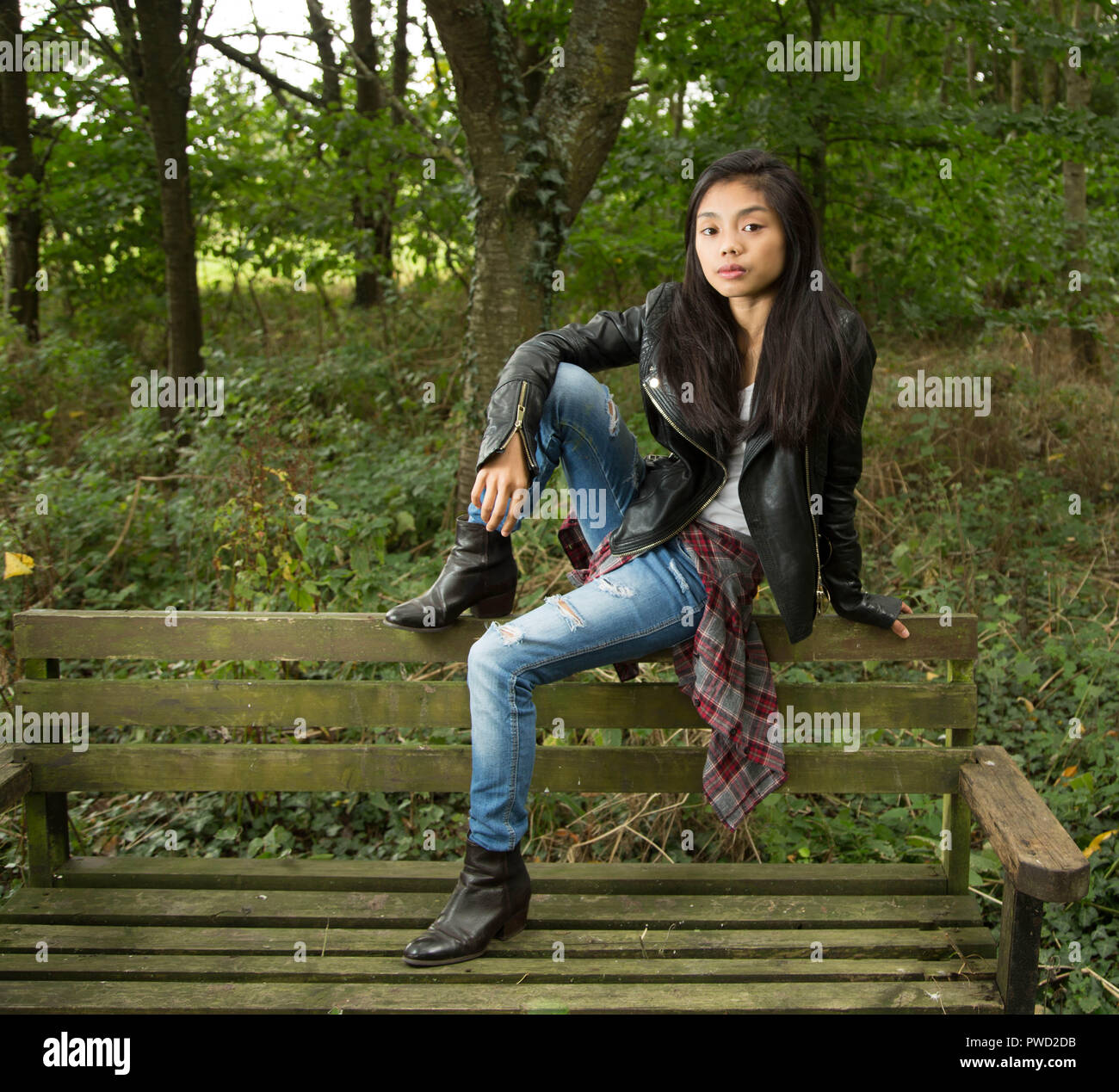 Young Filipino woman in black leather jacket in woods/forest setting sitting on old moss covered wooden bench in urban clothes. and jeans. Stock Photo