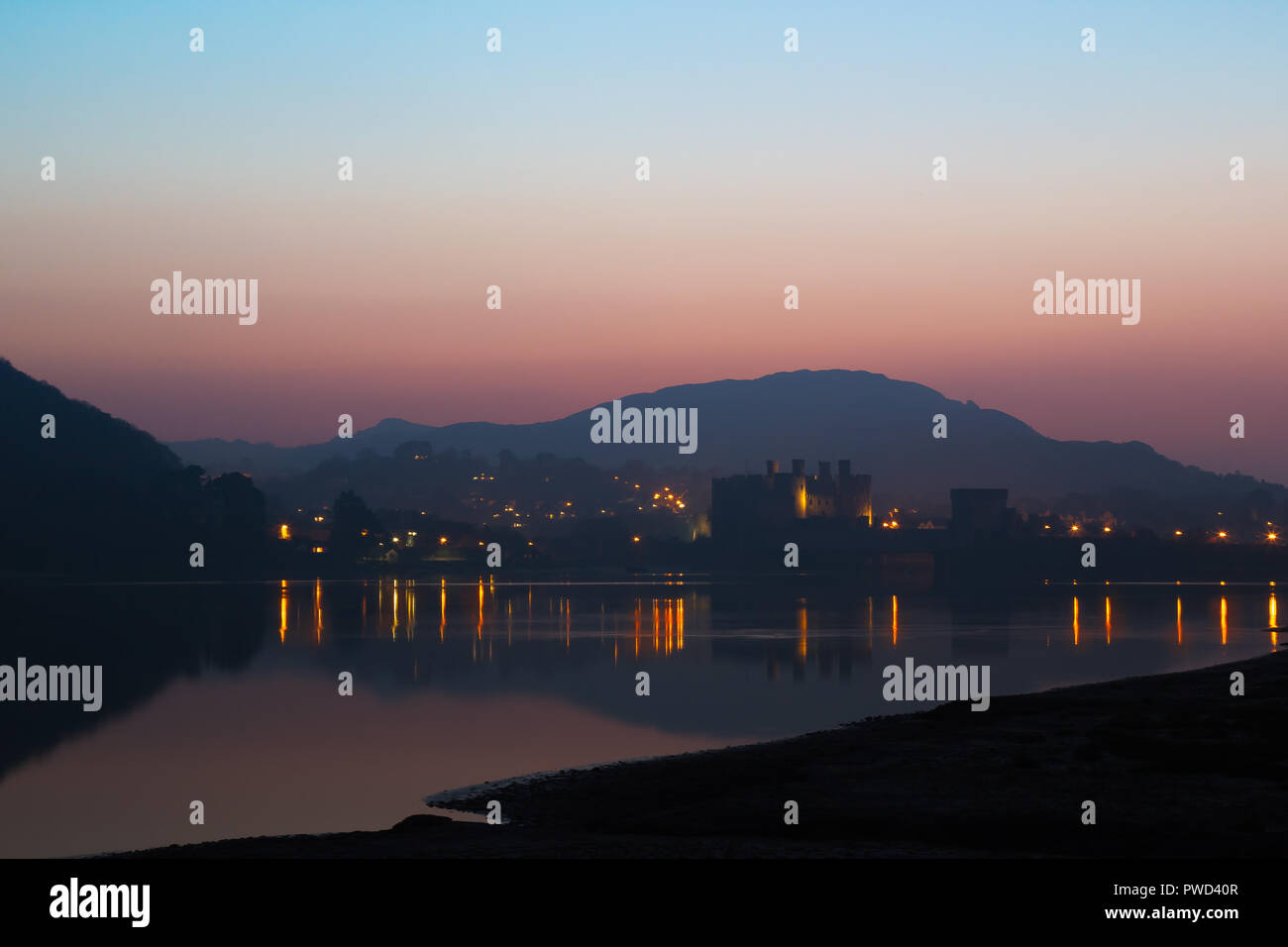 Conwy Castle lit up at night, almost in silhouette, picturesque scenic light reflections in harbour water. Dramatic sunset, twilight sky. Stock Photo