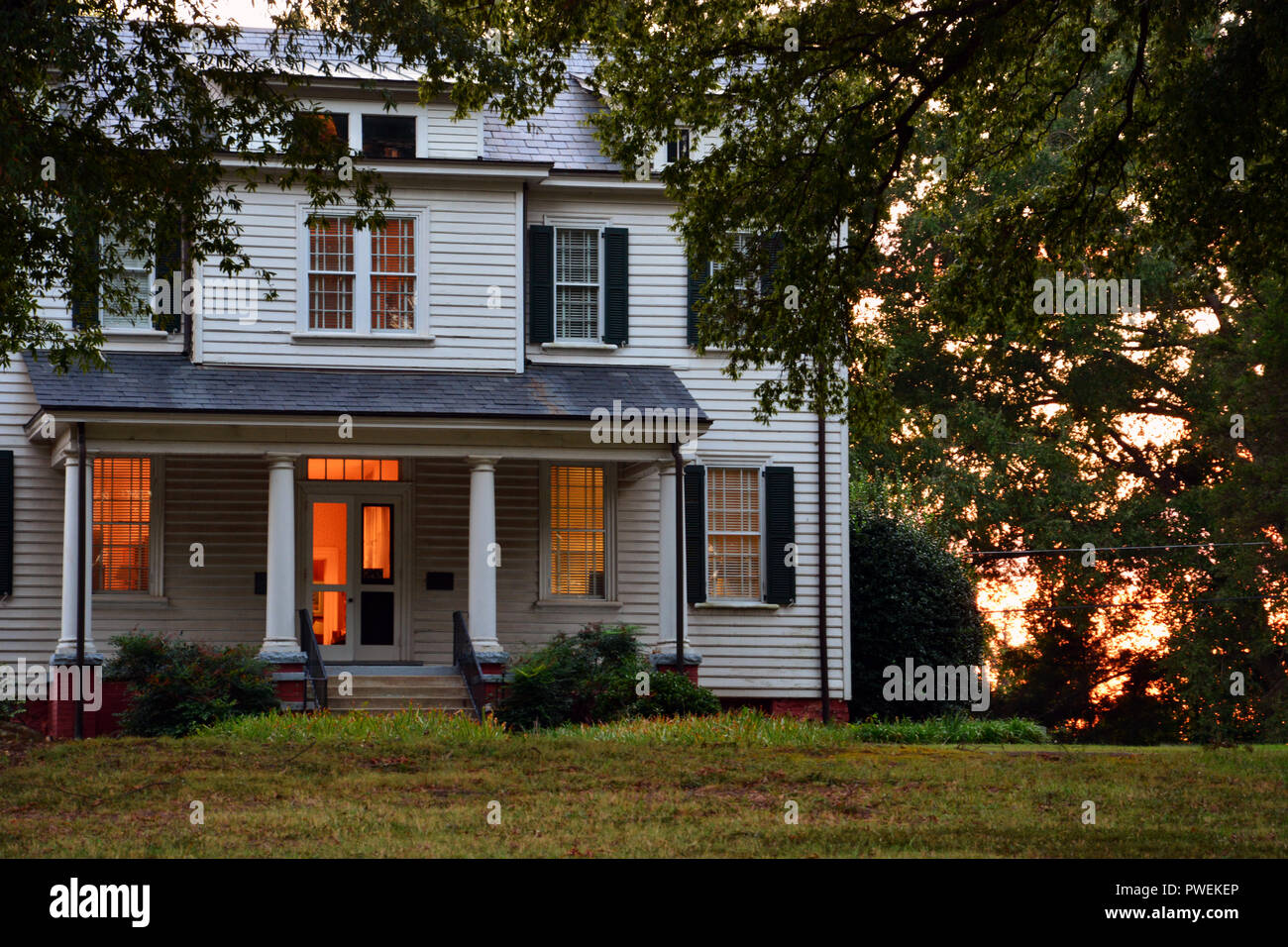 Lights create a warm glow inside a two story home in North Carolina as the sun sets behind the trees. Stock Photo