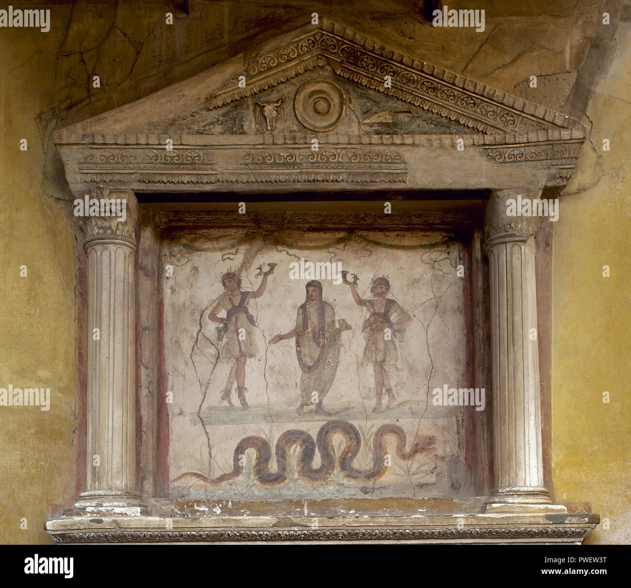 Pompeii. House of the Vettii. Lararium. Two Lares, holding a rhyton, flank a genius with a libation bowl and incense box, his head covered for a sacrifice. The snake, associated with the prosperity, approaches an altar. The tympanum shows a patera, an ox skull and a sacrificial knife. Italy. Stock Photo