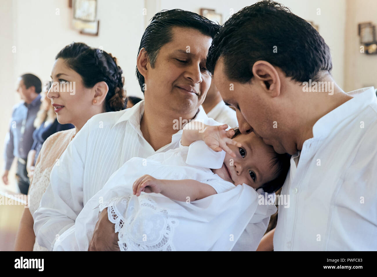 MERIDA,YUC/MEXICO - NOV 18, 2017: Godfather holds goddaughter during baptism ceremony at catholic church. Father kisses his baby girl. Stock Photo