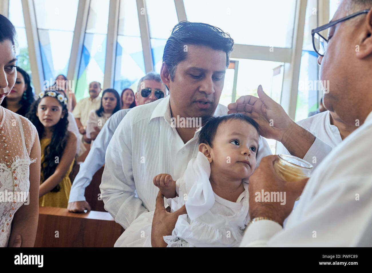 MERIDA, YUC/MEXICO - NOV 18, 2017: Godfather holds baby girl while catholic priest anoints her with chrism. Stock Photo