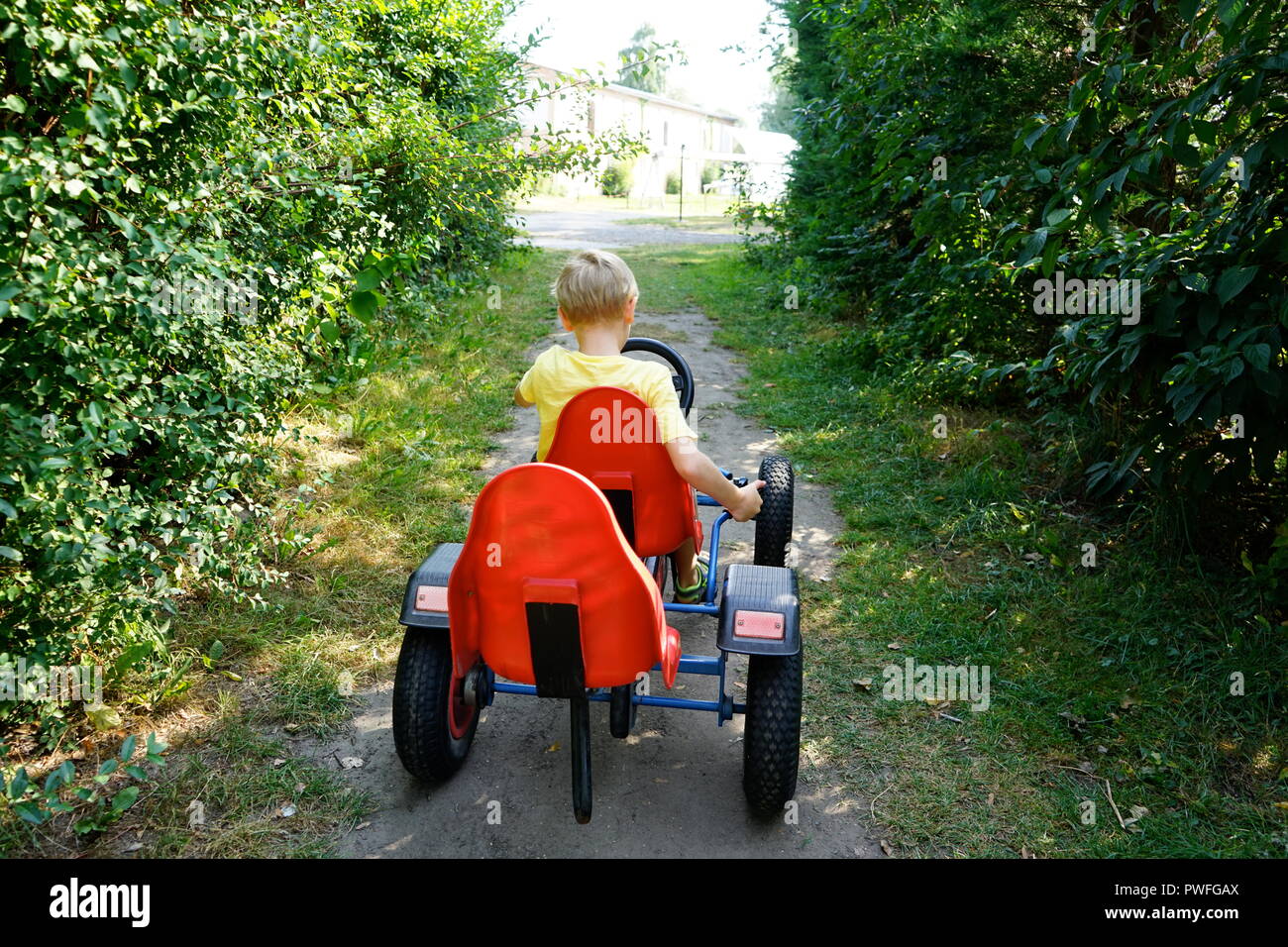 Active blond kid boy driving pedal toy car in green garden, outdoors. Stock Photo