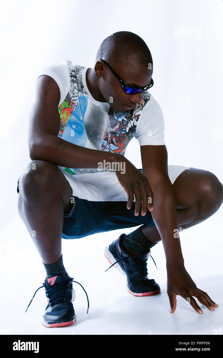 Young african male in a relaxed squatting position wearing sport clothes, sport shoes and sunglasses looking at his hand touching the ground Stock Photo