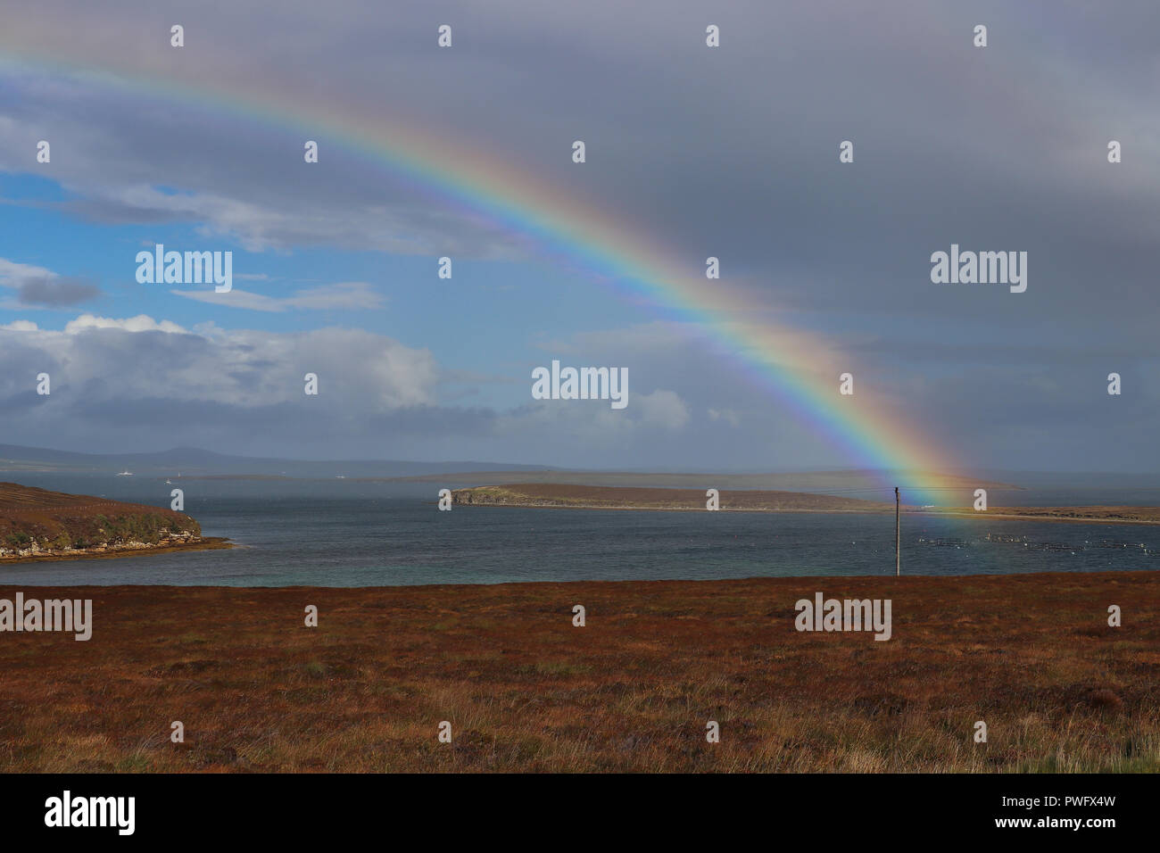 Gorgeous rainbow (double!) over Wide Firth, Orkney Mainland, Scotland, on a blustery, on and off rainy day. Blue sky, gray clouds, sea and brown field Stock Photo