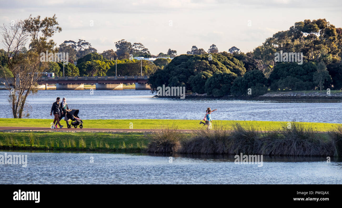Family parents walking with a stroller and children running ahead along the Swan River Perth Western Australia. Stock Photo