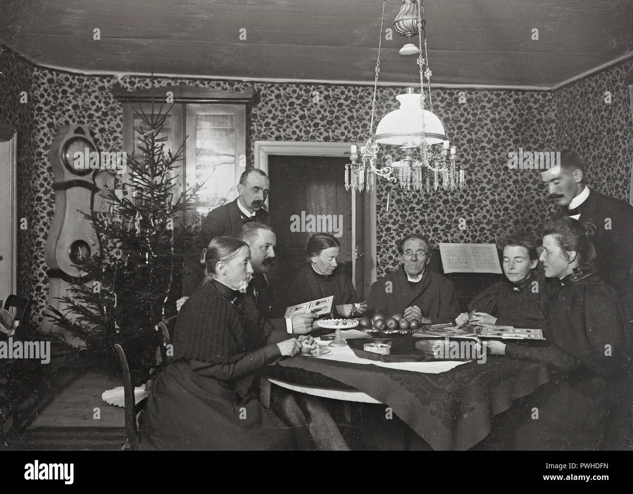 Christmas at the turn of the century 1800-1900. A priest has a central place at the table where the family is sitting. It's christmas holiday and they are enjoying the evening looking at cabinet photos of relatives and eating christmas apples. A christmas tree is standing in the corner. Sweden 1800s Ref 379A-13 Stock Photo