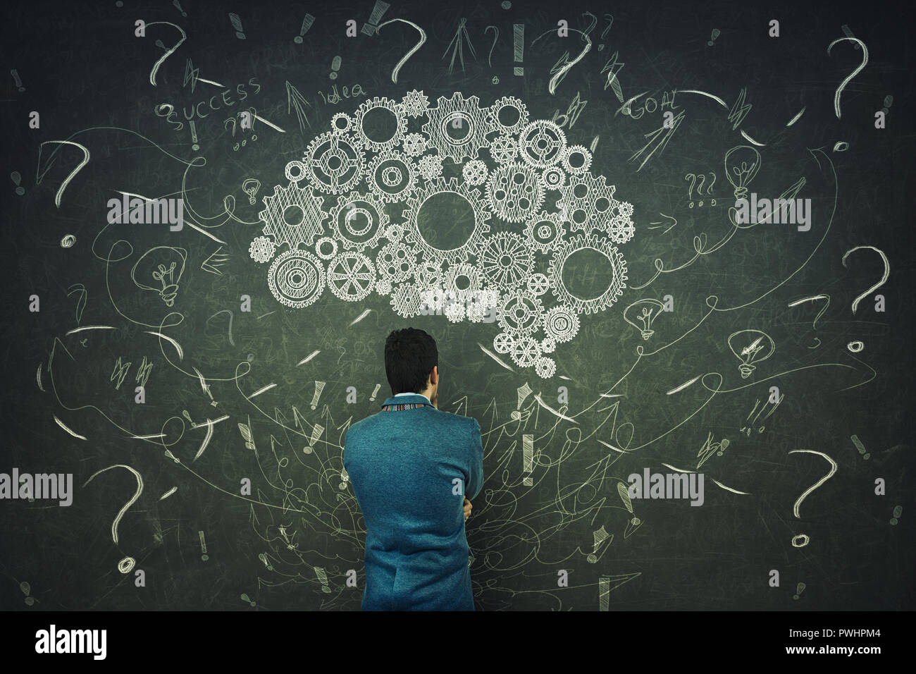 Rear view of a puzzled businessman having questions in front of a blackboard. Big chalk drawn gear brain above head, positive thinking mess as thought Stock Photo