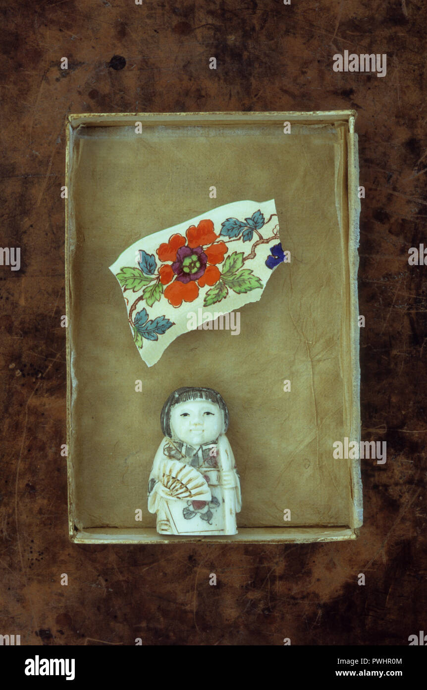 Pale netsuke of Japanese woman in traditional dress with fan lying in box with broken piece of flower pottery Stock Photo