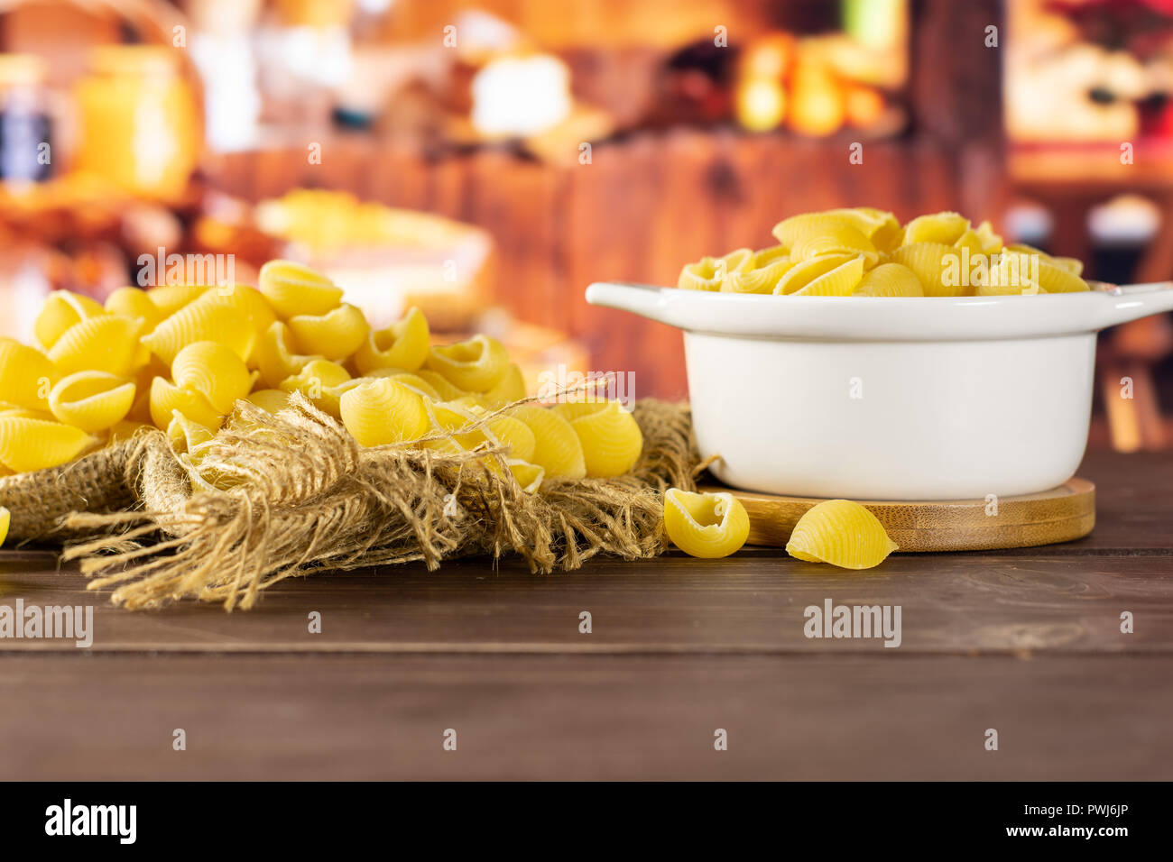 Lot of whole raw yellow pasta conchiglie variety on jute cloth in a ceramic stewpan with rustic wood kitchen in background Stock Photo