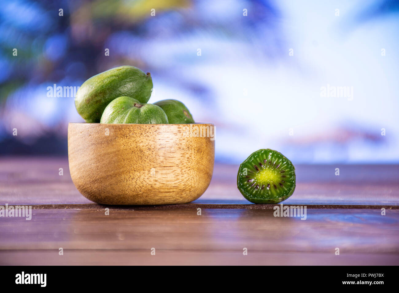 Lot of whole one half of fresh green mini baby kiwi fruit with wooden bowl with palm trees on the beach in background Stock Photo