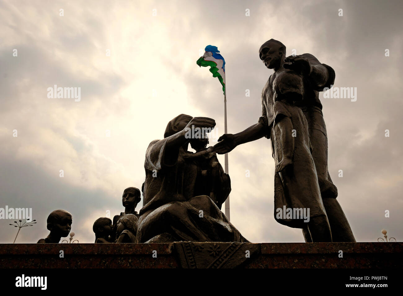 The Uzbek national flag flutters over a Soviet-era monument (created by sculptor Dmitry Ryabichev in 1982) to the blacksmith Shaakhmed Shamakhmudov and his wife Bahri Akramova who adopted 15 children of different nationalities, who lost their parents and were evacuated to Tashkent during WWII placed at the Park of Amity in Tashkent, capital of Uzbekistan. During Second World War more than one million evacuees, including more than 200,000 children who had lost their parents, were moved to Uzbekistan. Every day, between 200 and 400 orphans arrived at Tashkent railway station. Stock Photo