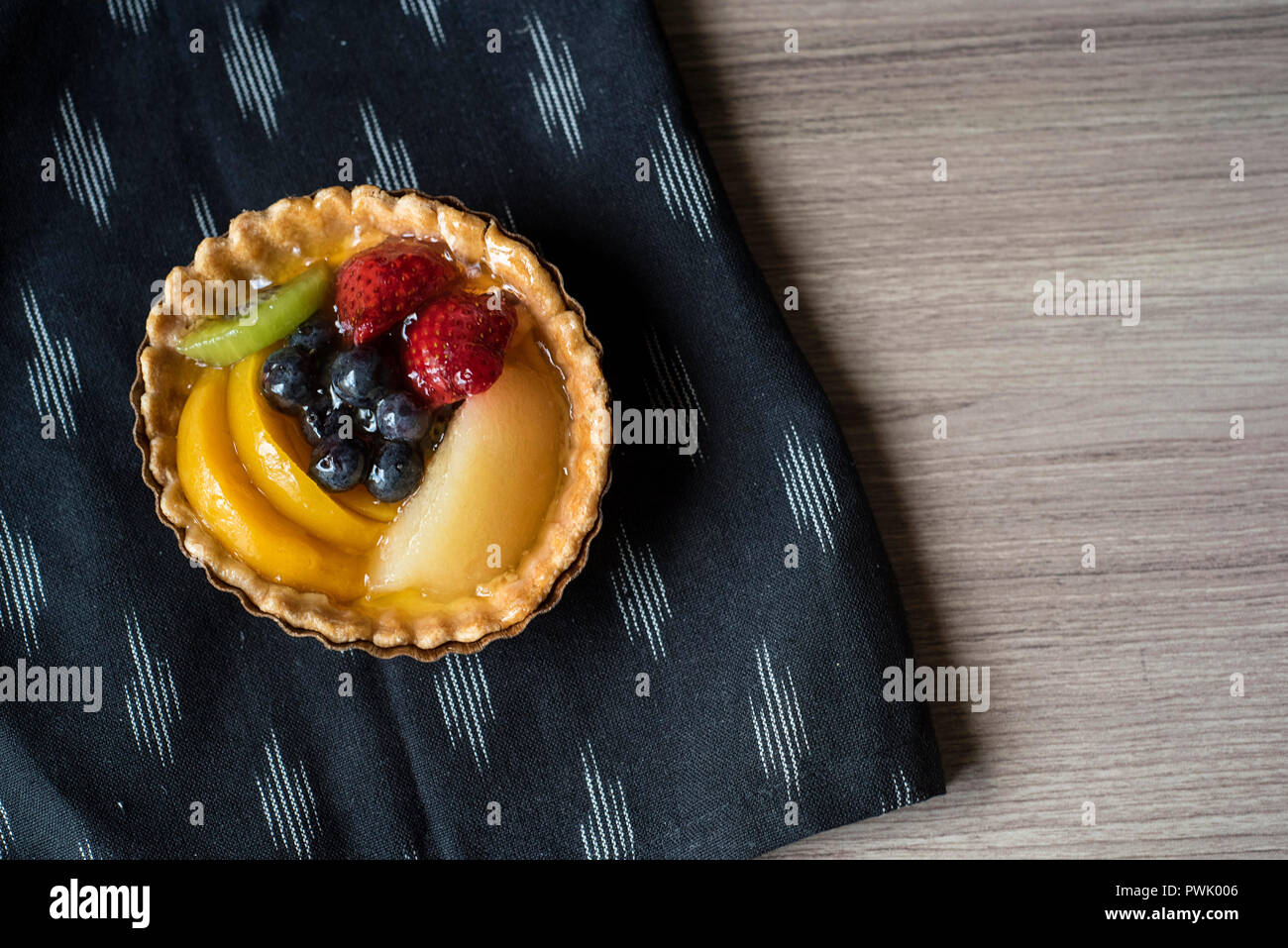 Overhead View of a Mixed Fruit Tart Individual Serving on a Patterned Napkin that rests on a Wood Counter Stock Photo