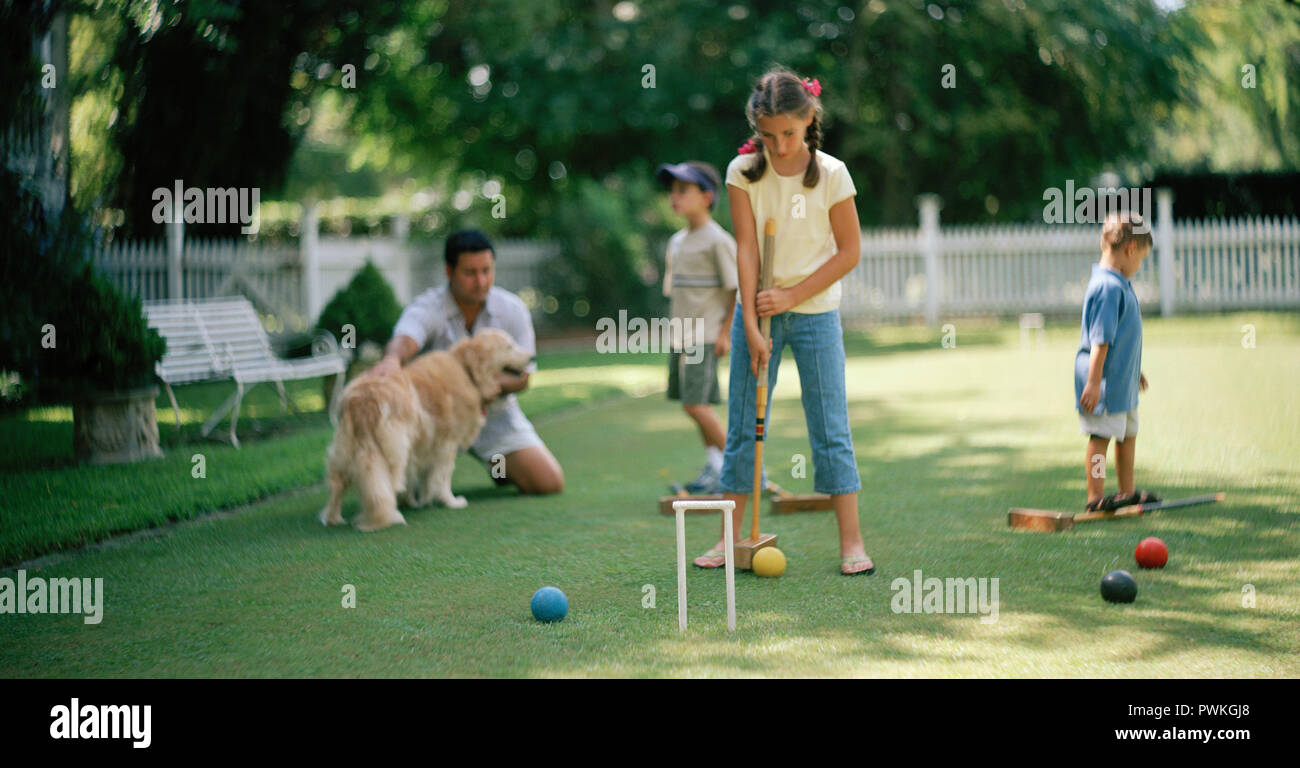 Girl playing croquet on a grassy lawn with her father and two younger brothers. Stock Photo