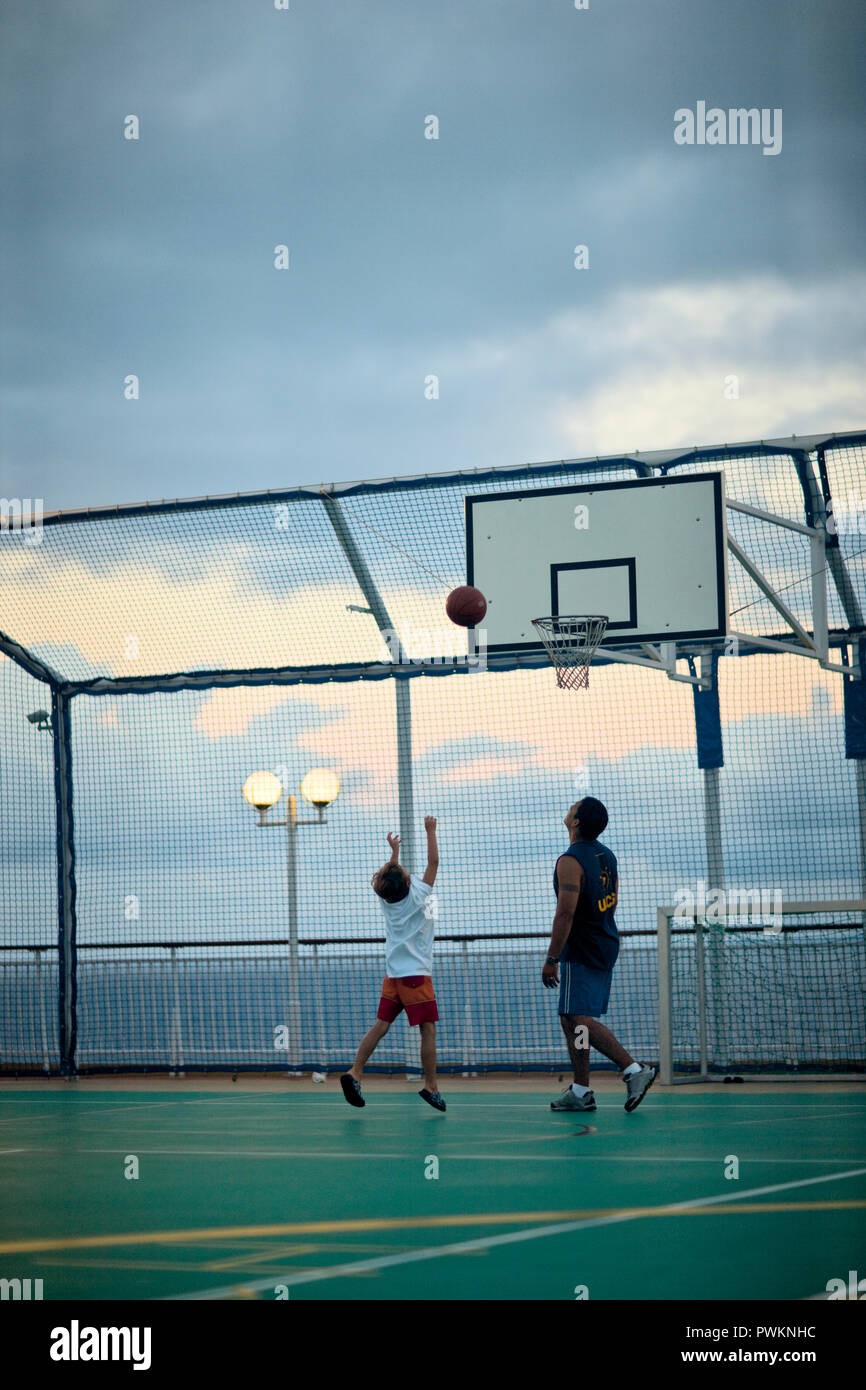 Man watches as a boy throws a basketball at a the hoop on a fenced seaside basketball court. Stock Photo