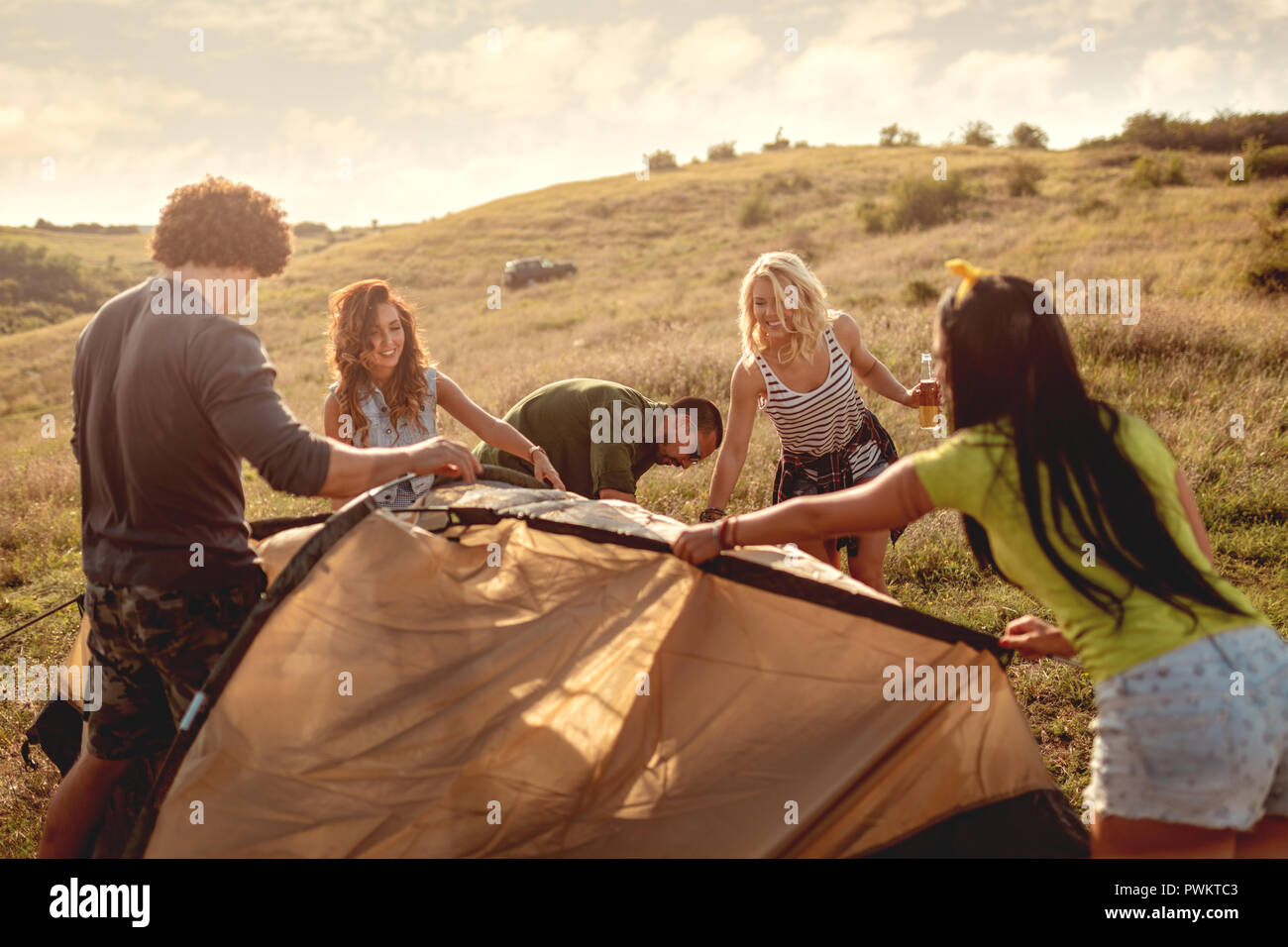 The young happy friends are preparing for camping. They're installing a tent on a suitable place in a meadow and their girlfriends are helping them. Stock Photo