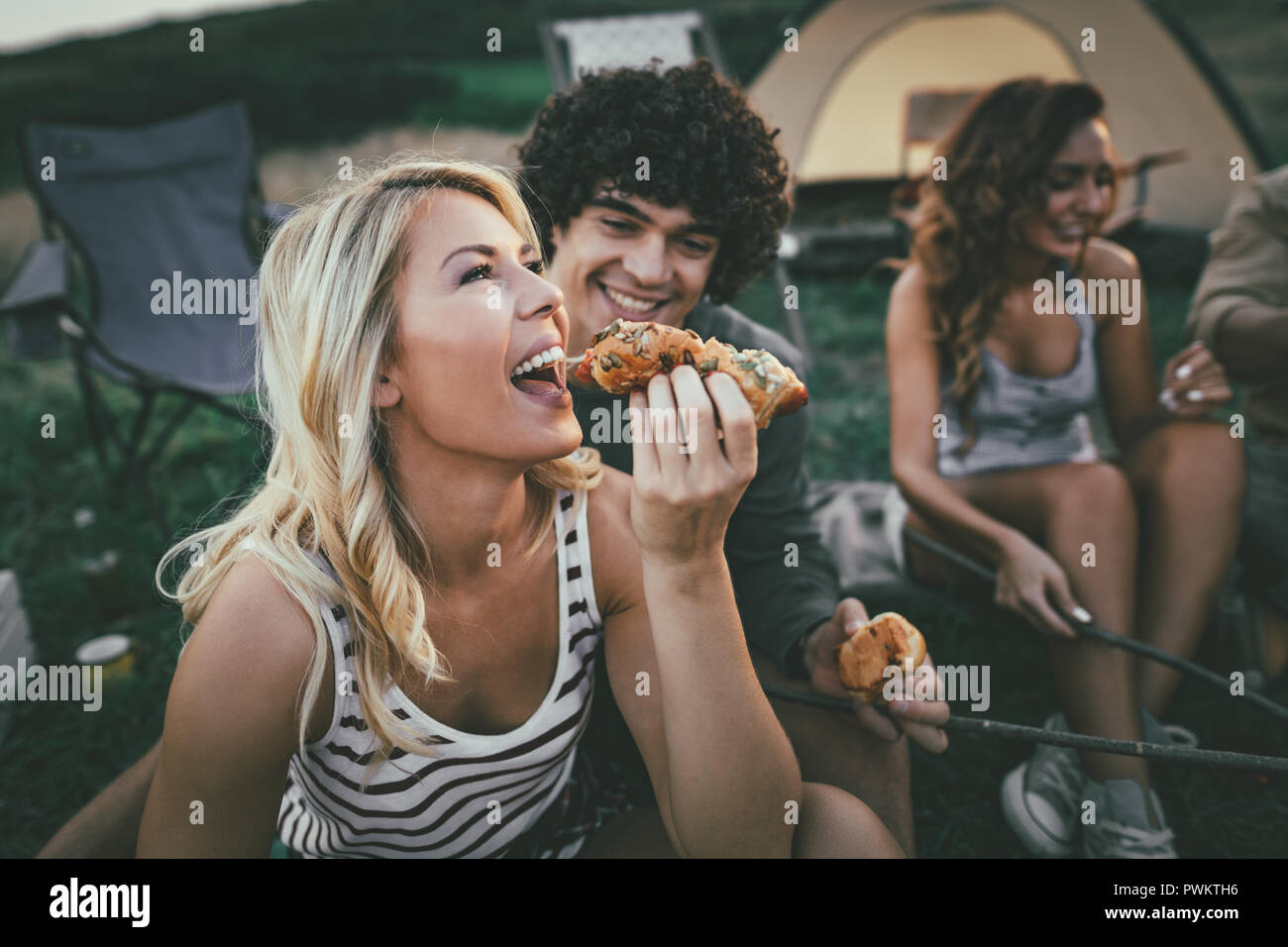 Happy young couple enjoy a sunny day with friends at the mountain. They're laughing and eating grilled sausages near tent. Stock Photo