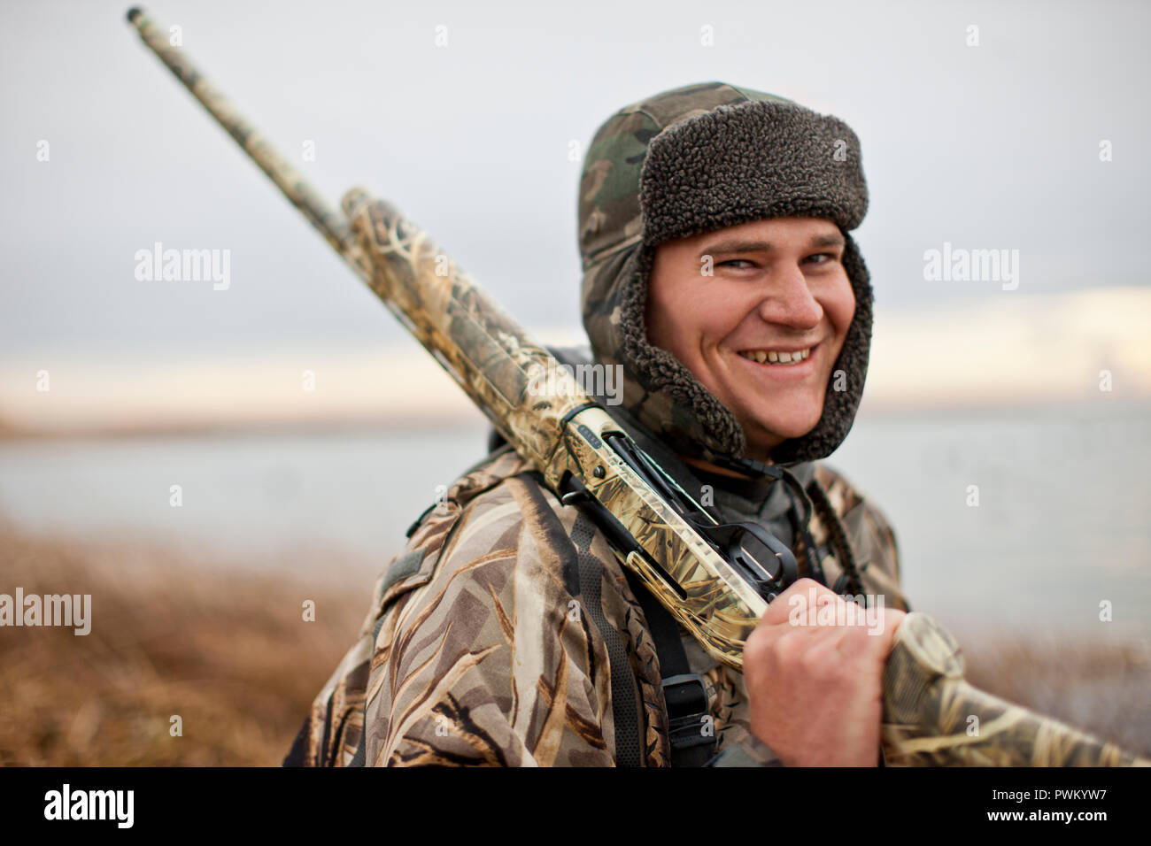 Portrait of a smiling duck hunter holding a shotgun while hunting dressed in camouflage clothing a rural lake. Stock Photo