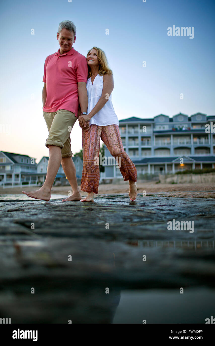 Mature couple holding hands and smiling as they walk barefoot on the beach at sunset. Stock Photo