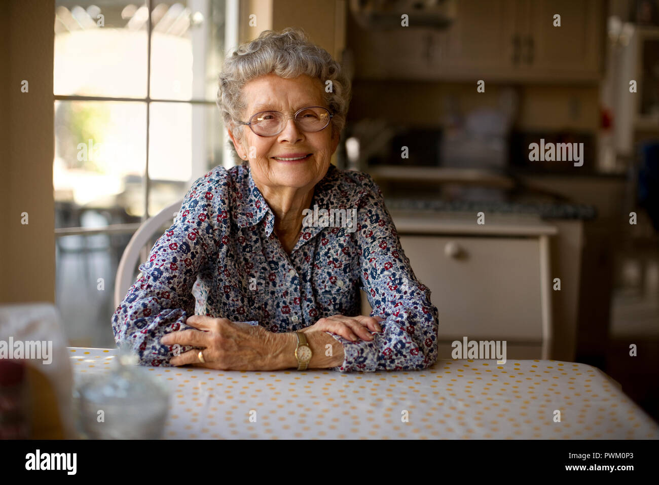 Portrait of a cheerful elderly woman sitting alone at her kitchen table ...