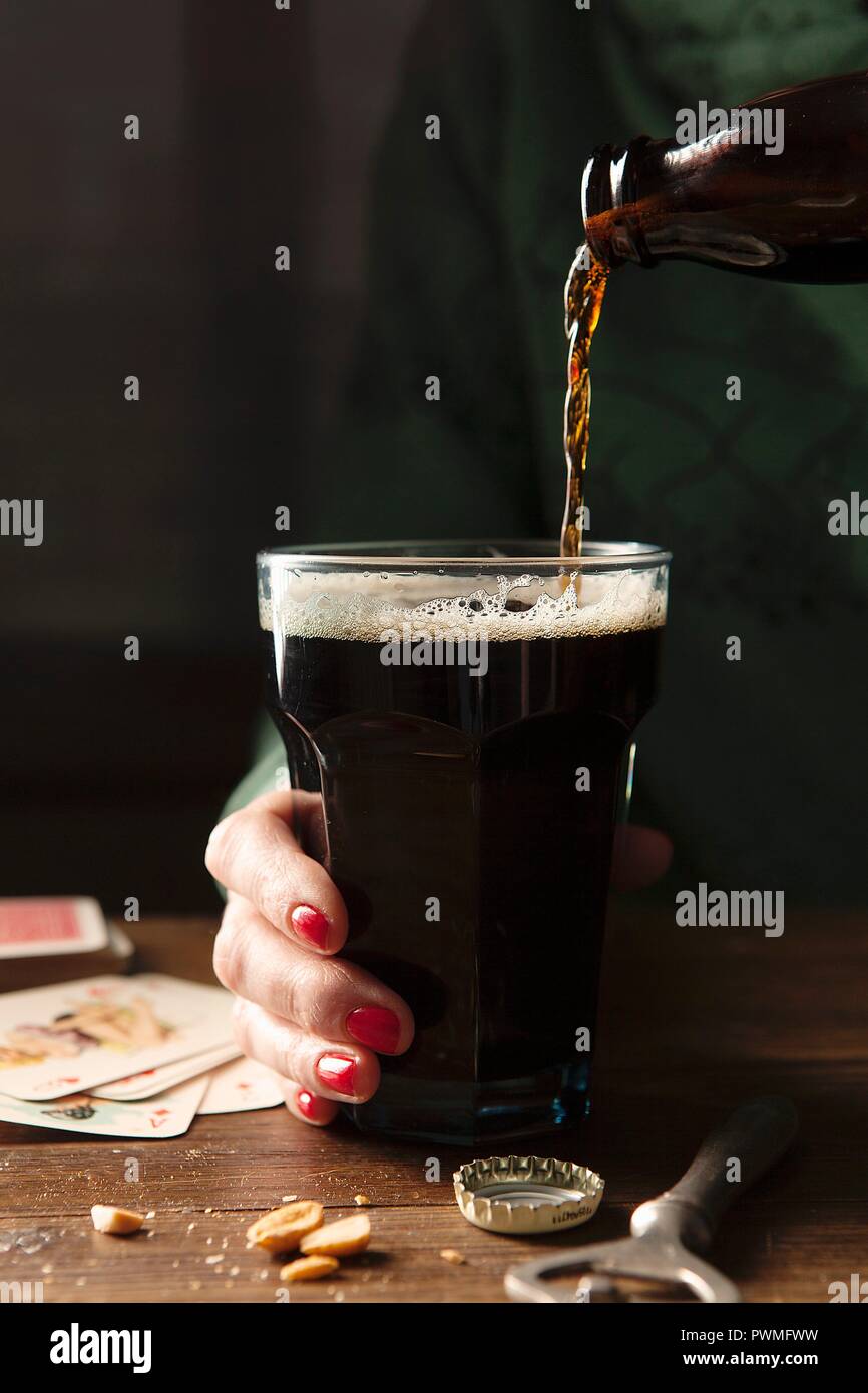 Bottle of Guinness being poured into a large glass being held by a hand with red nail varnish on a wooden table surrounds by the bottle top, bottle op Stock Photo