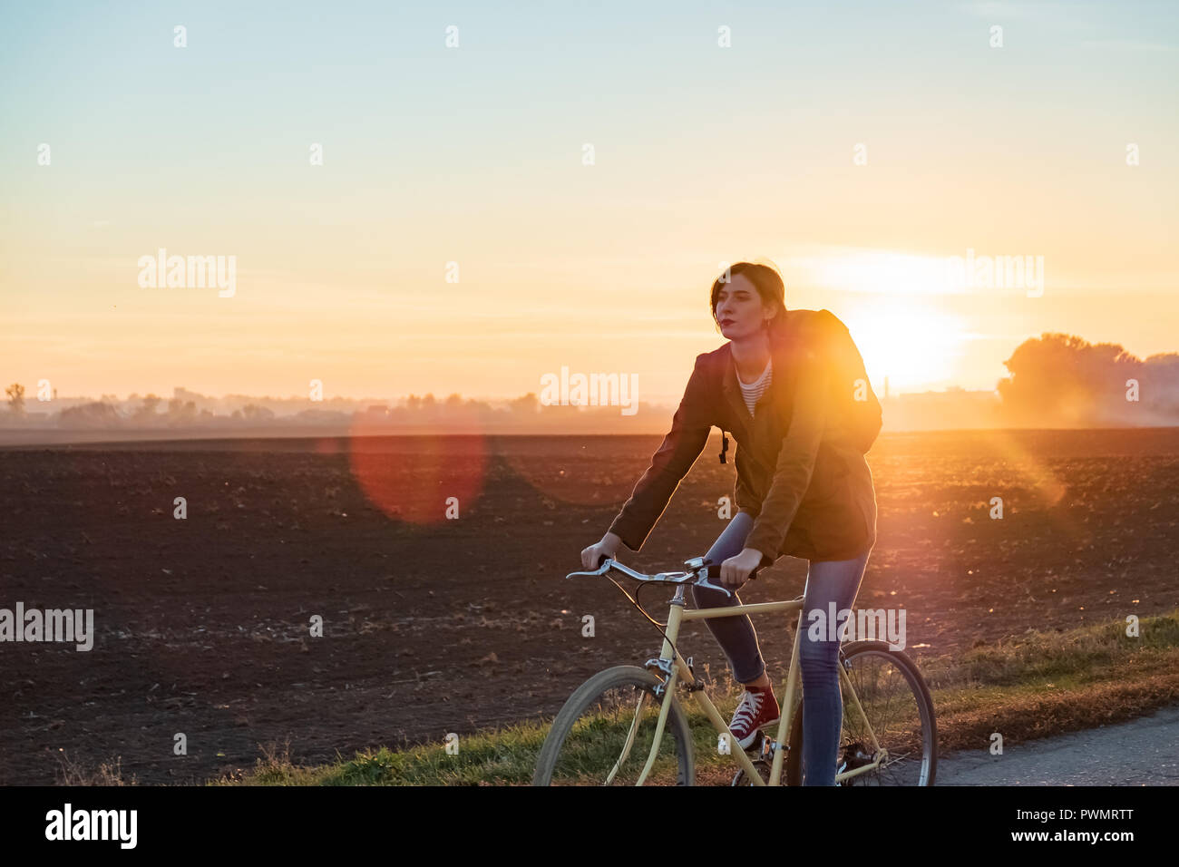 Female commuter riding a bike out of town. Woman cycling along the road at sunset Stock Photo