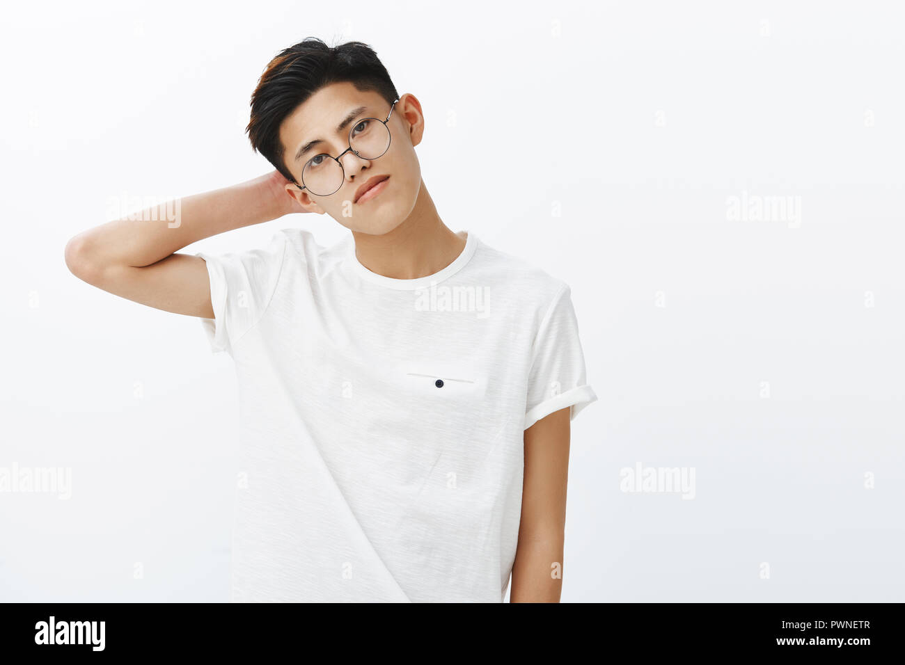 Attractive self-assured young asian with stylish hairstyle in glasses and white t-shirt tilting head touching back of neck making fashion pose gazing seriously at camera, dreaming become fashion model Stock Photo