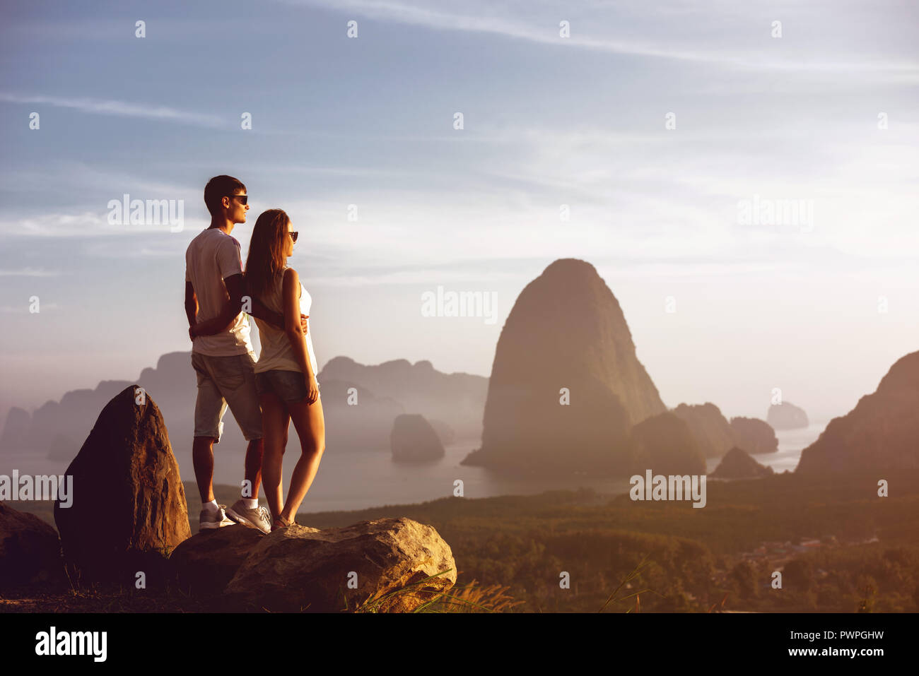 Young couple stands on big rock and looks at sunset or sunrise over sea bay and islands Stock Photo