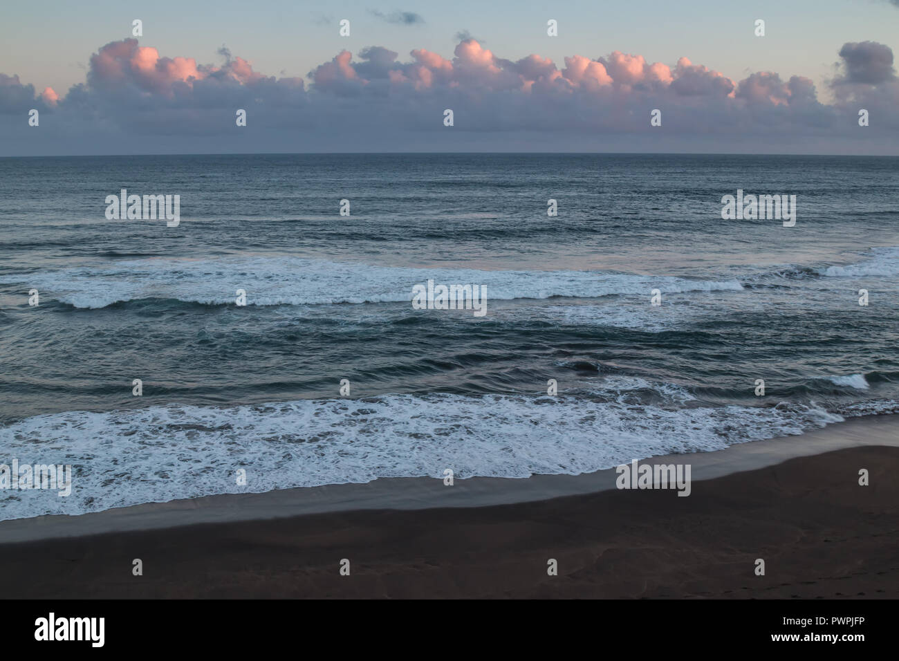 Small waves of the Atlantic ocean with its horizon. Dark sand of the beach in the foreground. Cloudy twilight partly pink clouds. Ponta Delgada, Sao M Stock Photo