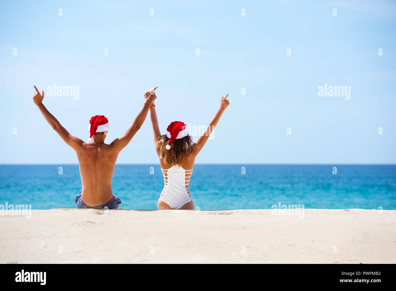 Happy couple sits at white sandy beach in christmas hats with raised arms. Sea vacations concept with place for text Stock Photo