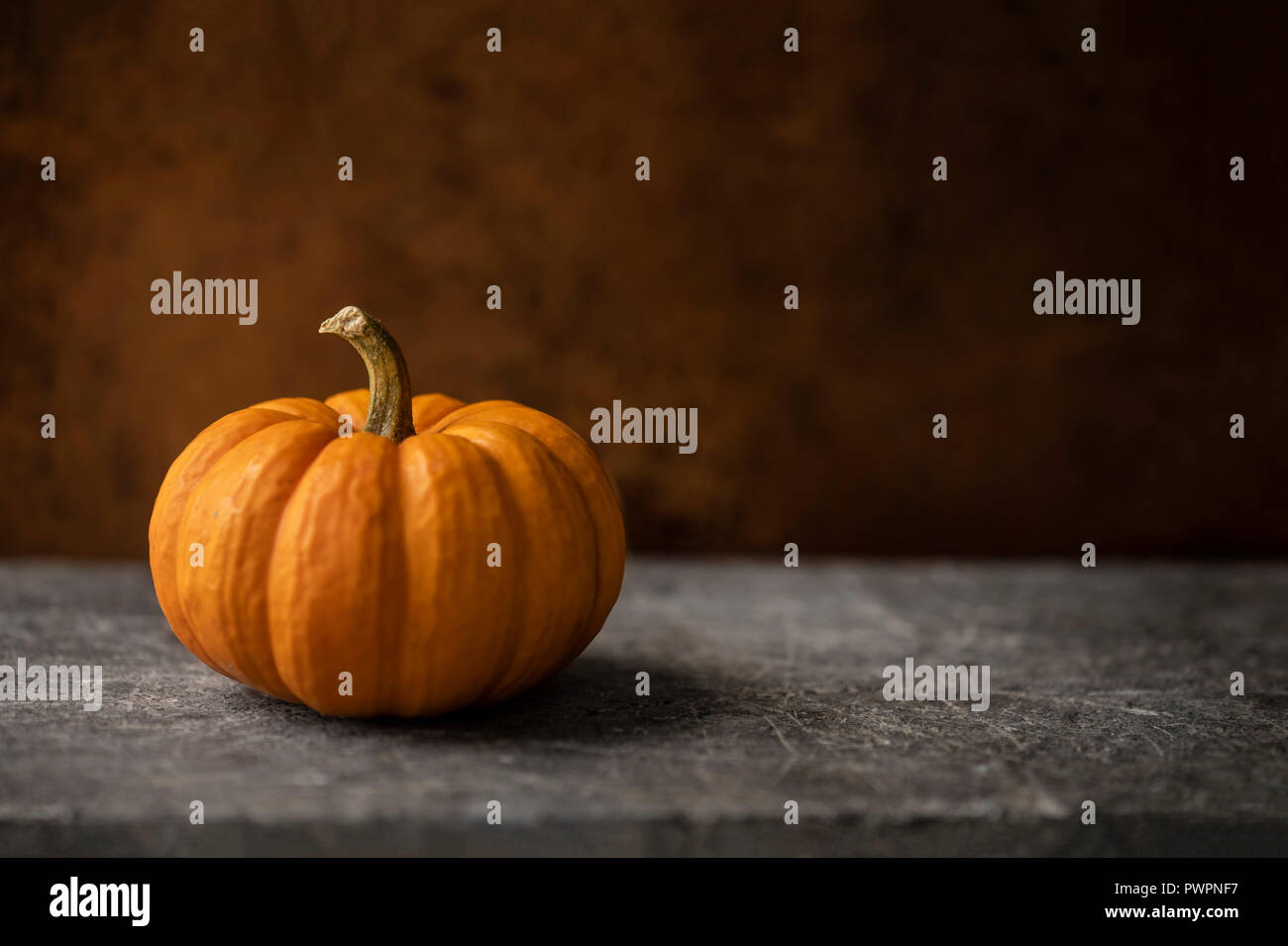 Mini orange pumpkin on a warn stone surface with rustic, brown, leather background. Moody natural lighting. Stock Photo