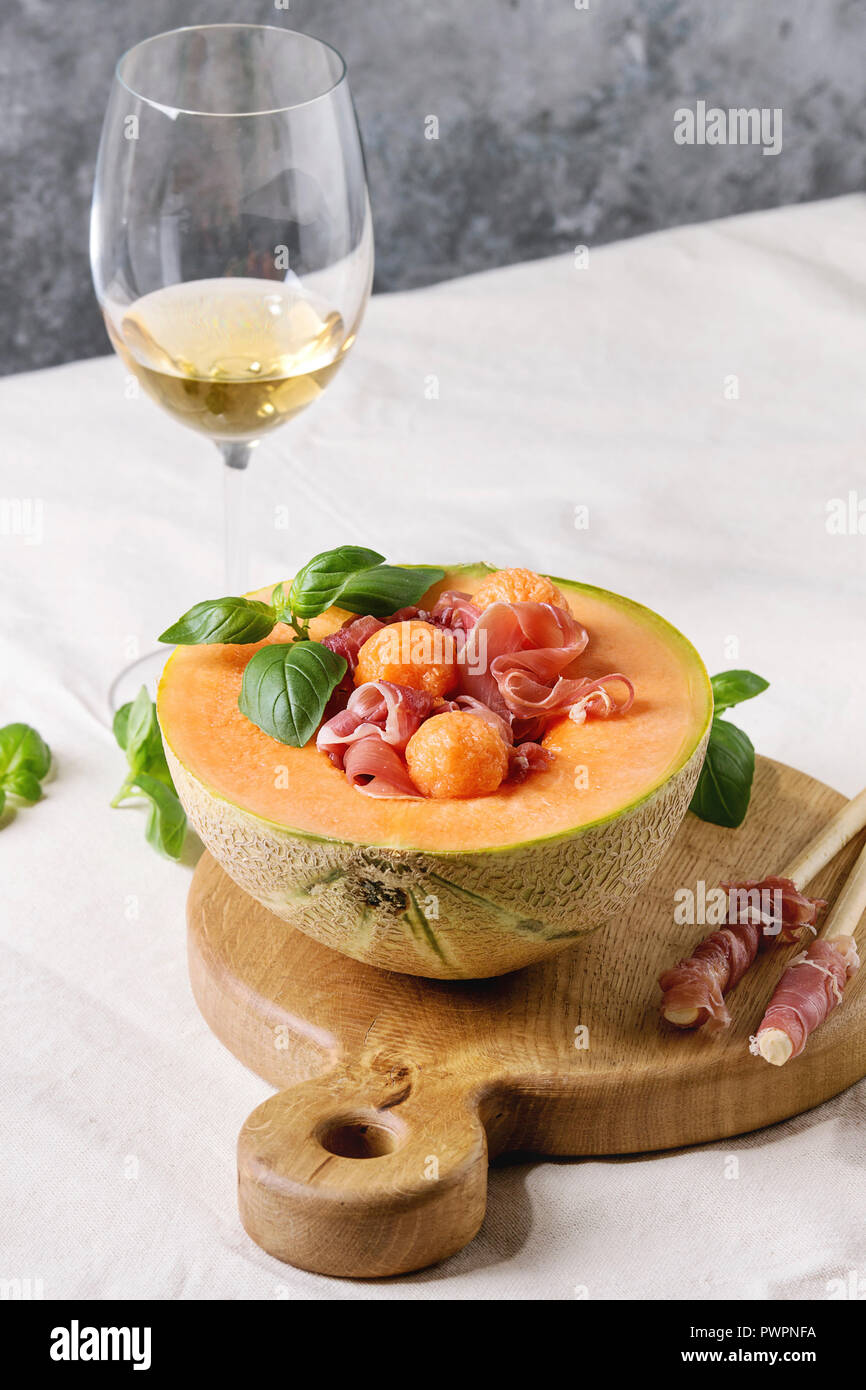 Melon and ham or prosciutto salad served in half of Cantaloupe melon, decorated by fresh basil standing on wooden serving board over white tablecloth Stock Photo