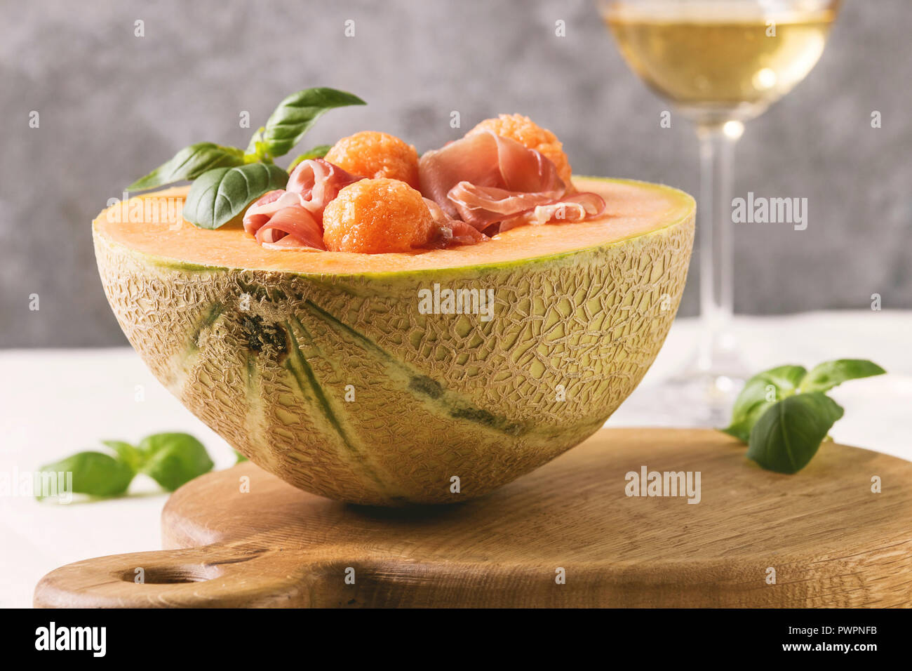 Melon and ham or prosciutto salad served in half of Cantaloupe melon, decorated by fresh basil standing on wooden serving board over white tablecloth Stock Photo