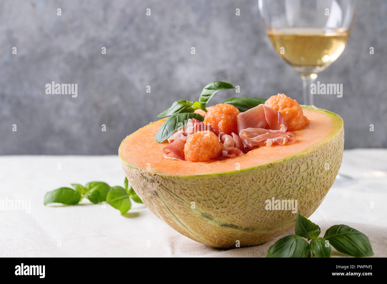 Melon and ham or prosciutto salad served in half of Cantaloupe melon, decorated by fresh basil standing on white tablecloth with glass of white wine. Stock Photo