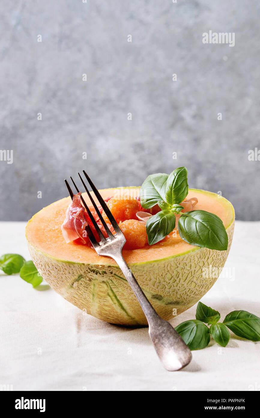 Melon and ham or prosciutto salad served in half of Cantaloupe melon, decorated by fresh basil standing on white tablecloth with fork. Stock Photo