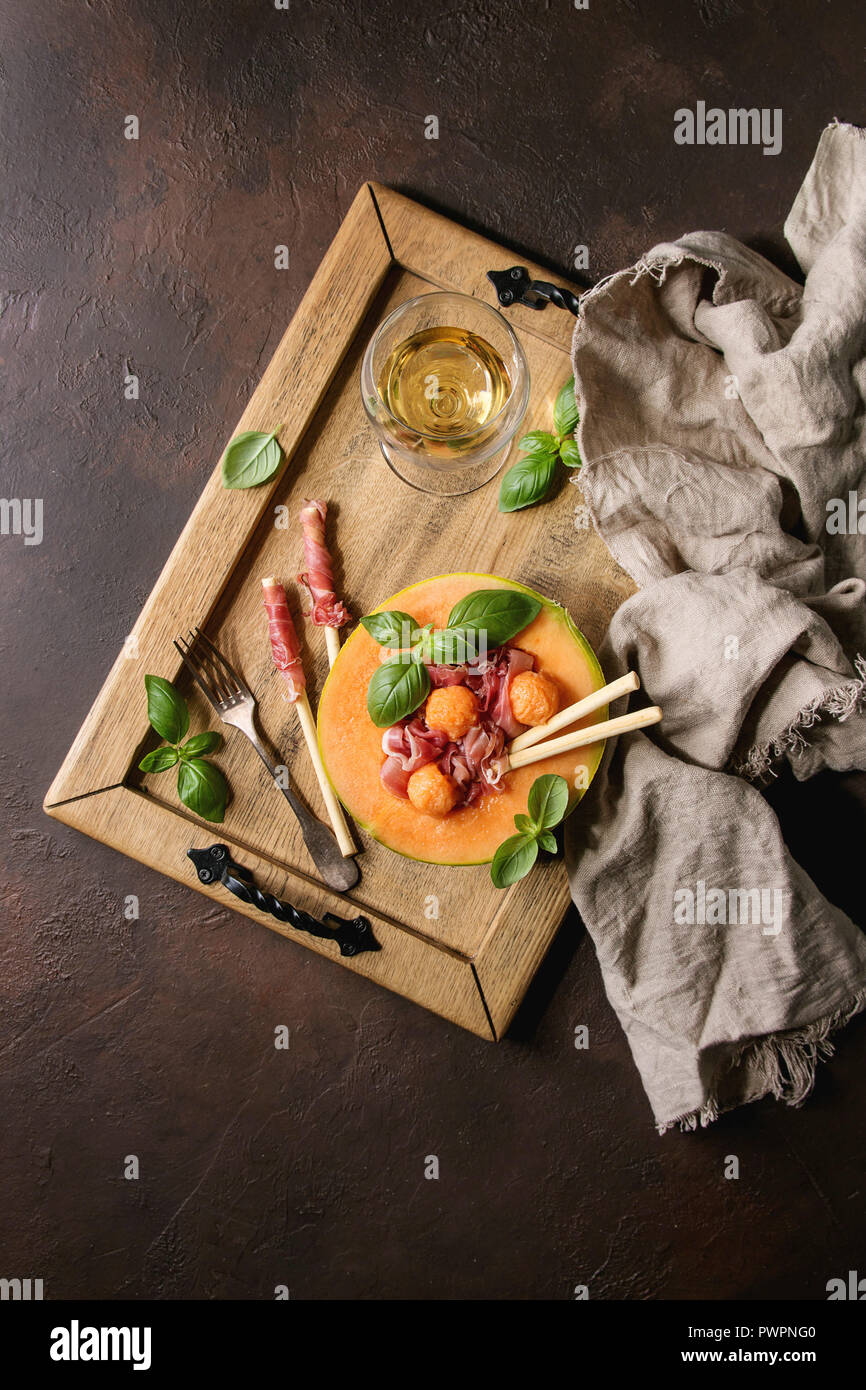 Melon and ham salad served in half of Cantaloupe melon with fresh basil and grissini bread on wooden serving tray over dark brown texture background w Stock Photo