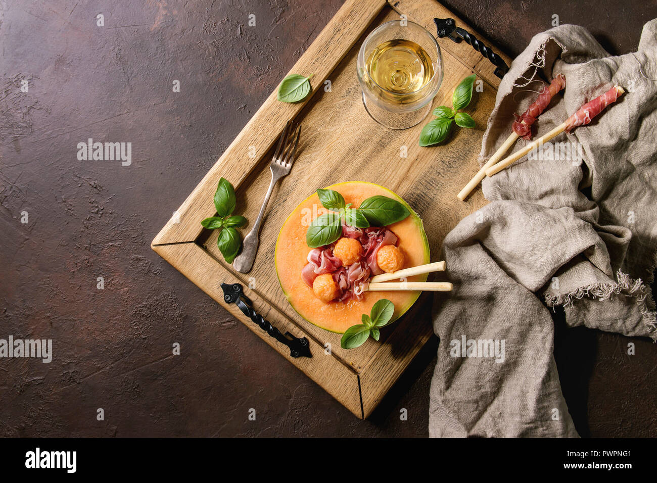 Melon and ham salad served in half of Cantaloupe melon with fresh basil and grissini bread on wooden serving tray over dark brown texture background w Stock Photo