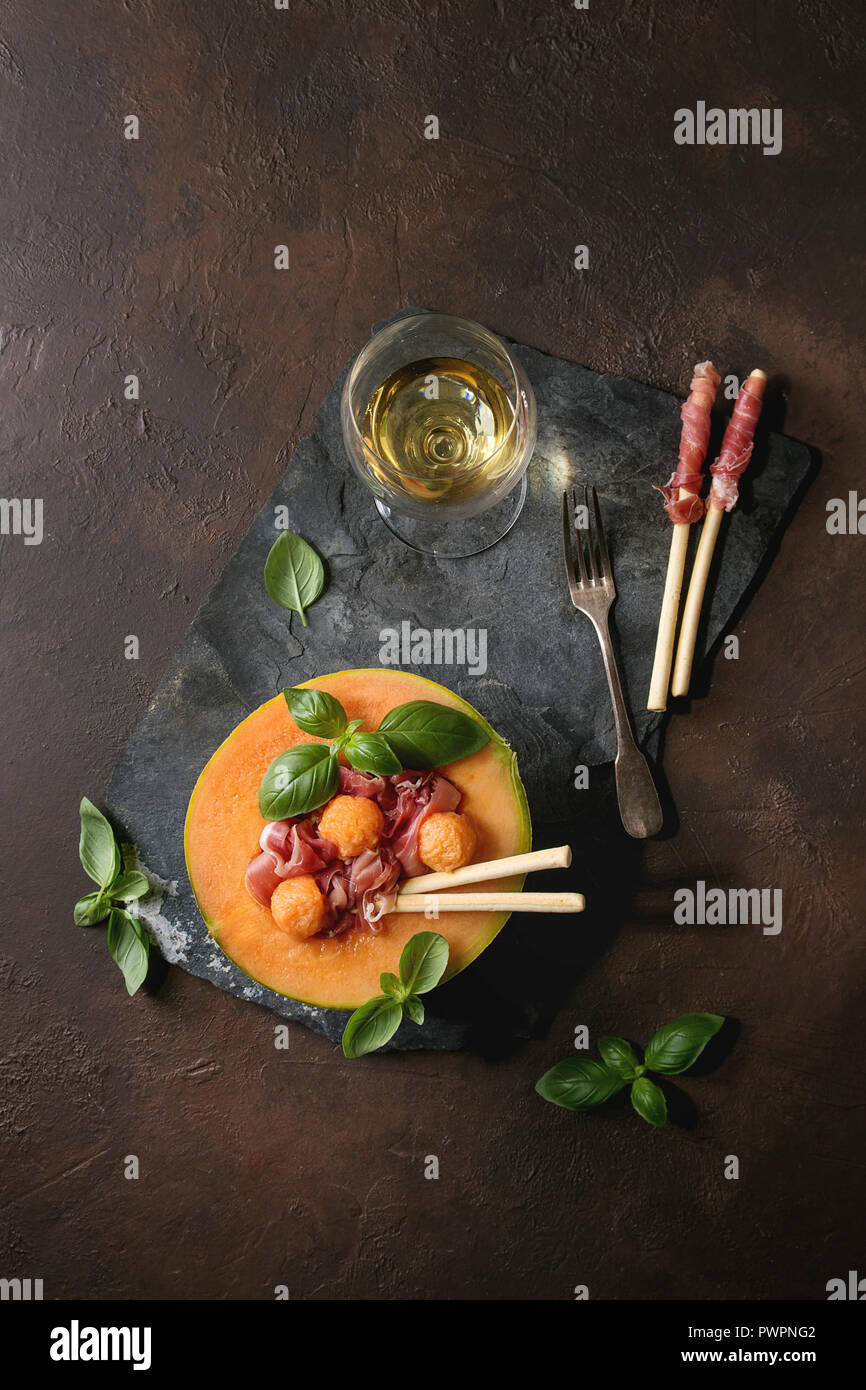 Melon and ham salad served in half of Cantaloupe melon with fresh basil and grissini bread on black slate board over dark brown texture background wit Stock Photo