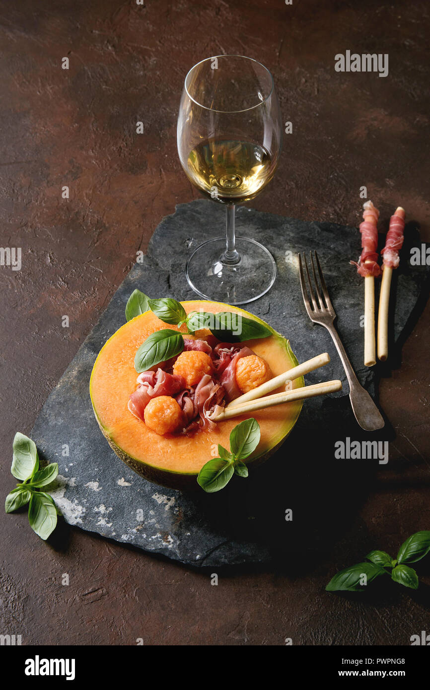 Melon and ham salad served in half of Cantaloupe melon with fresh basil and grissini bread on black slate board over dark brown texture background wit Stock Photo