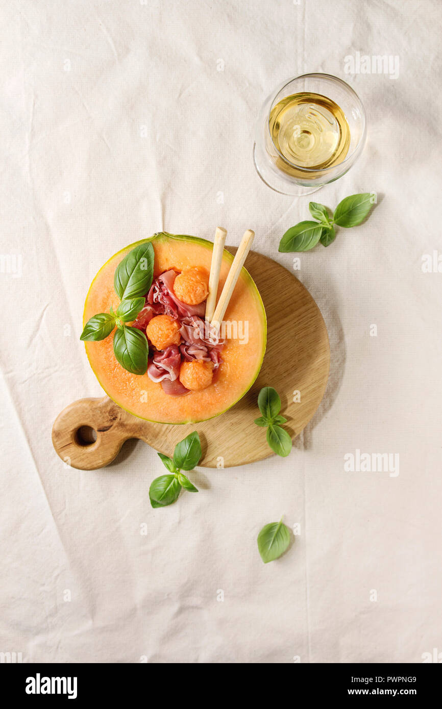 Melon and ham or prosciutto salad served in half of Cantaloupe melon, decorated fresh basil and grissini bread on wooden serving board over white line Stock Photo