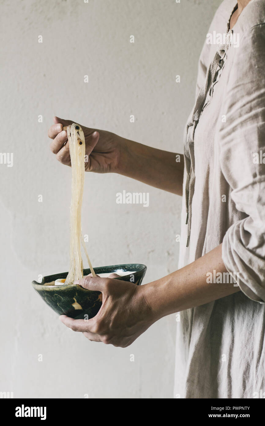 Woman in white dress holding in hands bowl of Asian rice Noodle Soup and take noodles by chopsticks. Grey wall as background. Asian style dinner. Stock Photo