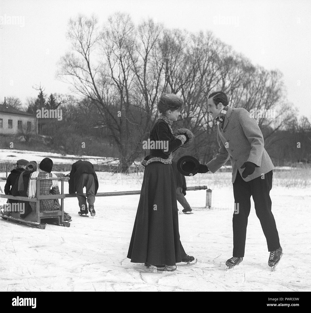 Figure skating at the turn of the century 1800-1900. A couple is pictured together when skating outdoors. In the background a typical swing sledge with children playing with it. Picture most probably taken on a film set. Sweden 1952. Photo Kristoffersson ref BF53-1 Stock Photo
