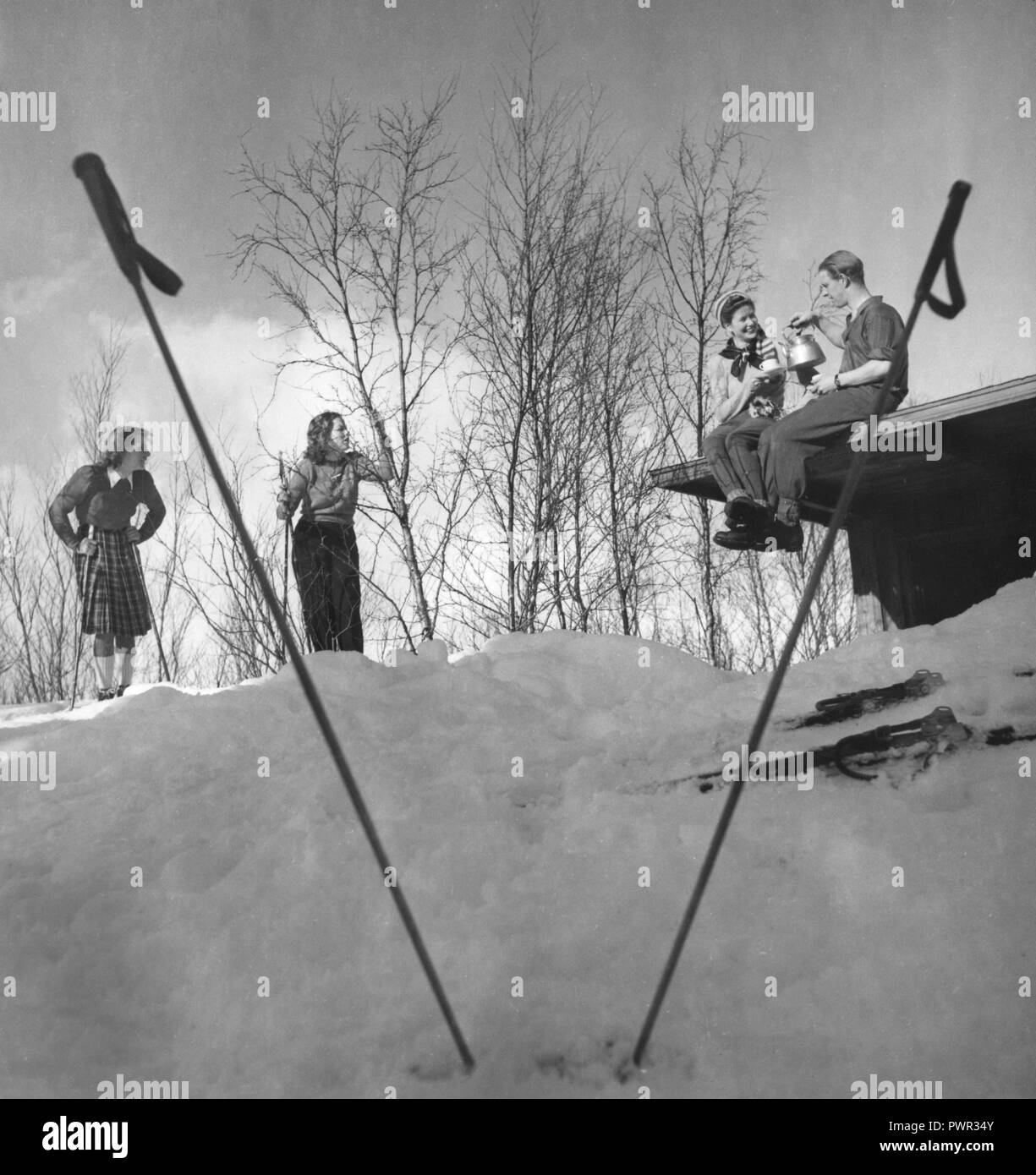 Winter in the 1940s. A young couple is having a cup of coffee while sitting on the roof of their cottage. Their two friends are standing on skis and chatting.  Sweden 1940s. Photo Kristoffersson Ref D115-2 Stock Photo