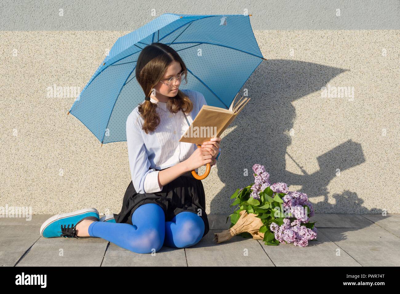Portrait of romantic teenage girl with bouquet of lilacs, with an umbrella reading book on gray wall background. Outdoor, copy space Stock Photo