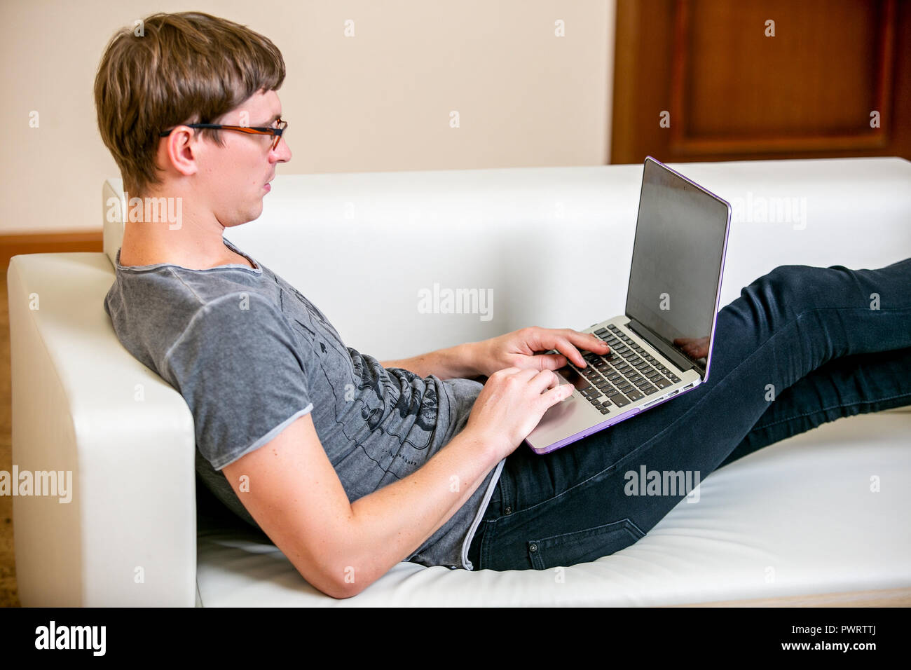 Concentrated young man with glasses working on a laptop in a home office. Prints on the keyboard and scrolls text on the screen while lying on the sofa Stock Photo