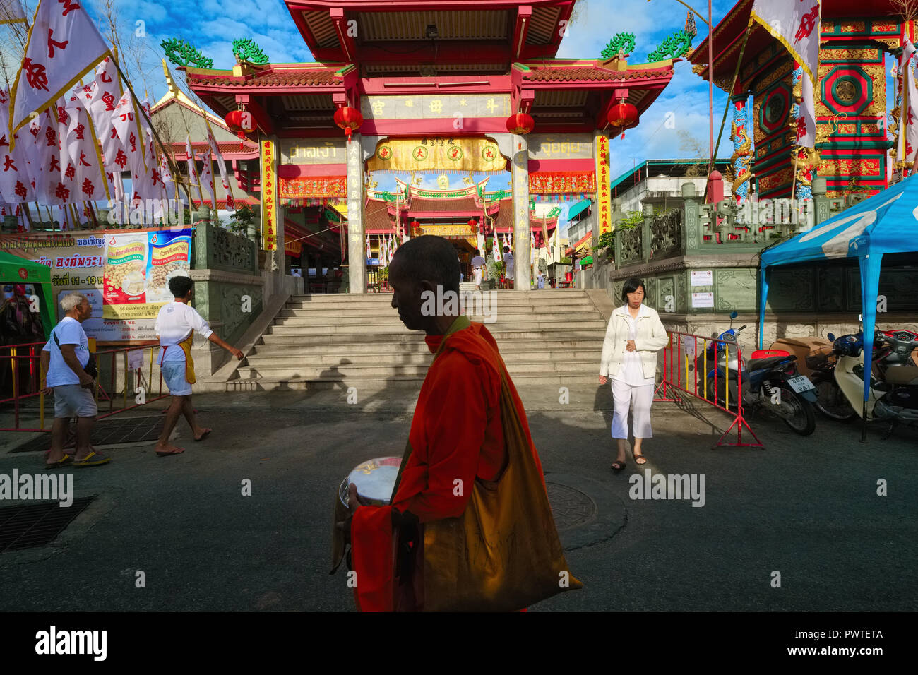 A Thai Buddhist monk passes the Taoist Jui Tui Temple in Phuket Town, Phuket, Thailand, on his early morning alms round, the alms bowl in his hands Stock Photo