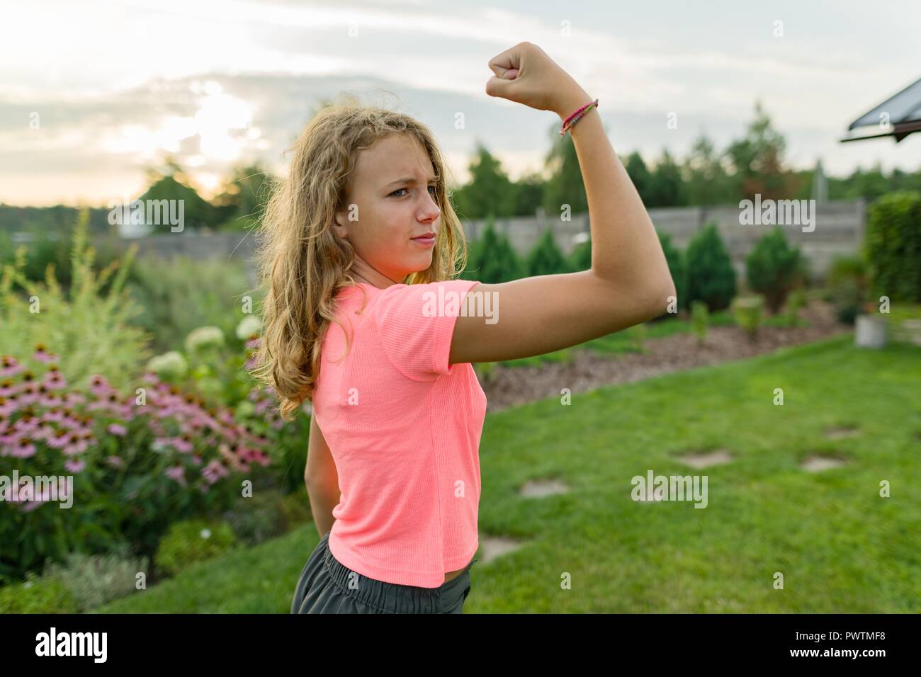 People, power, stamina, strength, health, sport, fitness concept. Outdoor portrait smiling teenage girl flexing her muscles, background green lawn sun Stock Photo