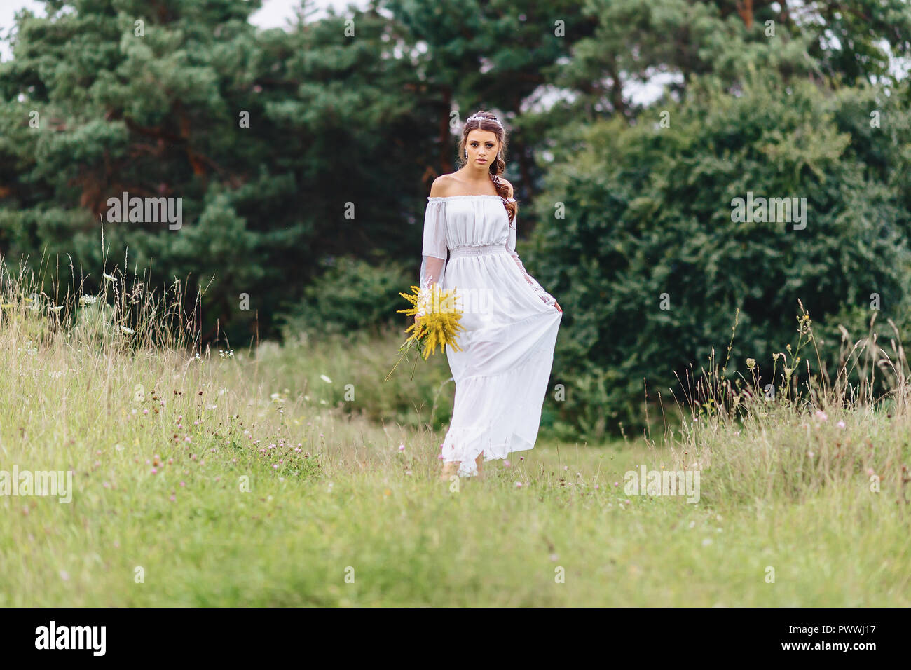 young pretty girl with flower in her hands in light dressing walk at lawn near green summer forest Stock Photo