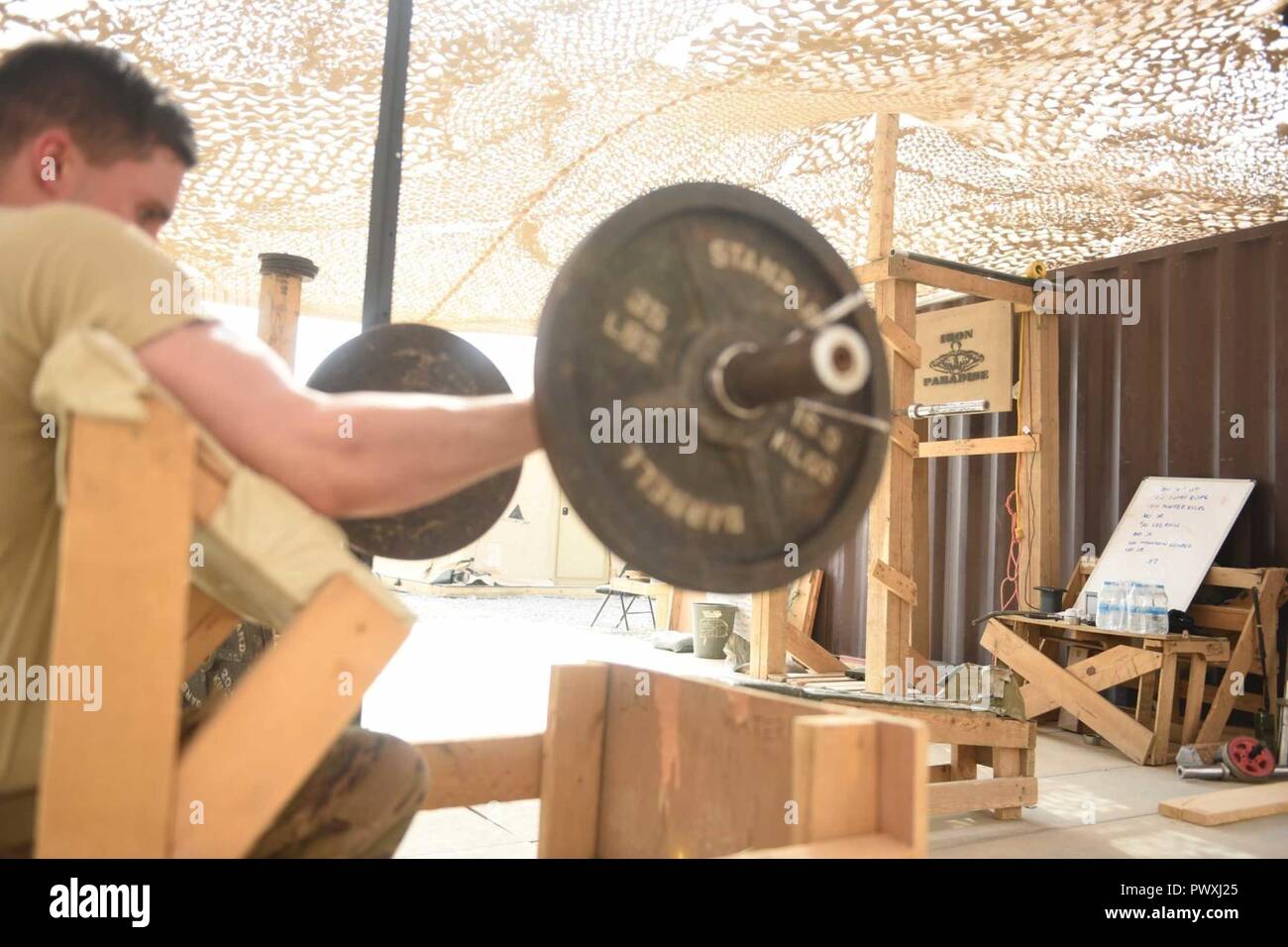 U.S. Air Force Senior Airman Justin Salisbery, an airfield management specialist deployed in support of Combined Joint Task Force – Operation Inherent Resolve and assigned to the 370th Air Expeditionary Advisory Group, Detachment 1, curls a barbell on a makeshift preacher curl bench in the 370th AEAG detachment’s “Iron Paradise” gym at Qayyarah West Airfield, Iraq, July 2, 2017. The air advisors built the “Iron Paradise” gym, which includes a makeshift squat rack, bench press, preacher curl bench and other recently acquired weights, creating an area often filled with U.S. Air Force and Army pe Stock Photo