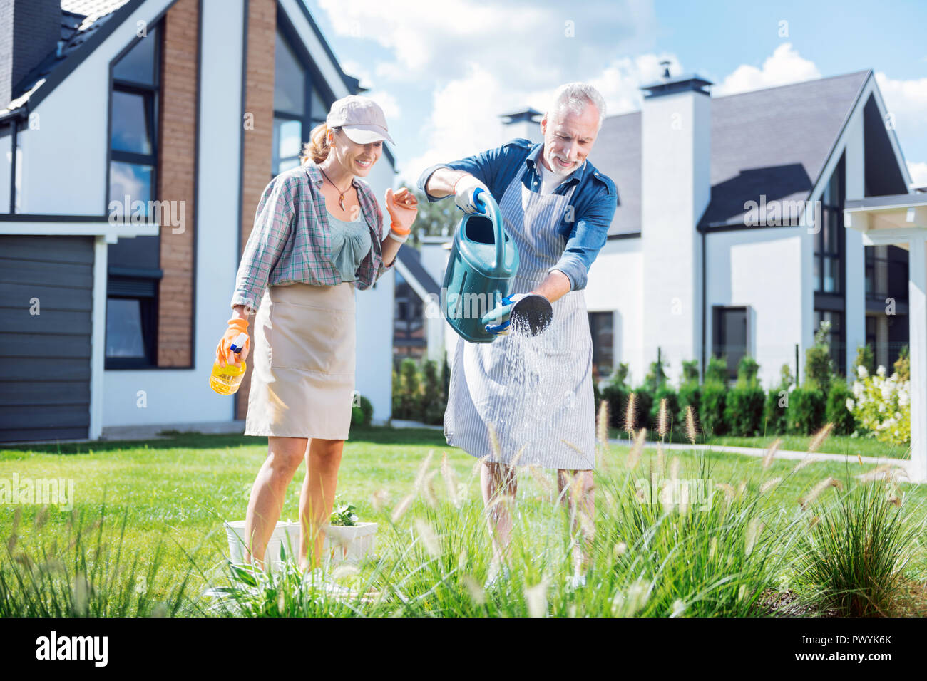 Bearded grey-haired man holding big garden sprinkler watering plants with wife Stock Photo